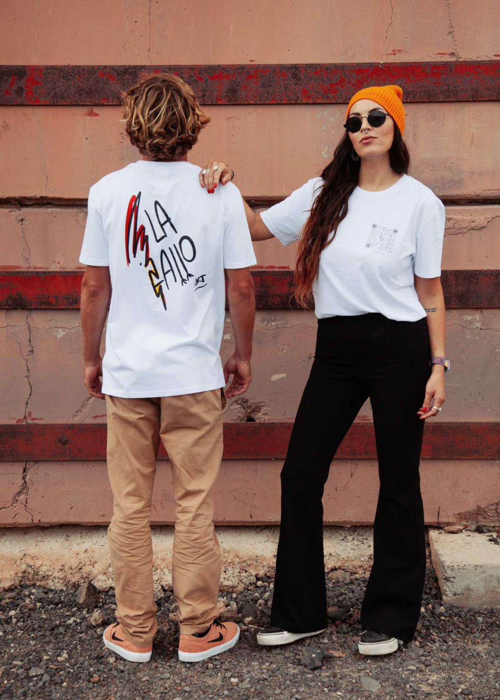 Pareja con camiseta “La Gallo Art” blanca, vista de espalda. Logo artístico de la marca impreso en algodón ecológico.